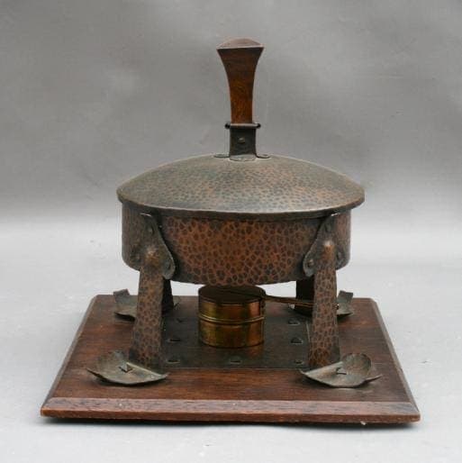 Antique metal Arts And Crafts Chafing Dish on a wooden base, featuring embossed designs and a central handle on the lid.