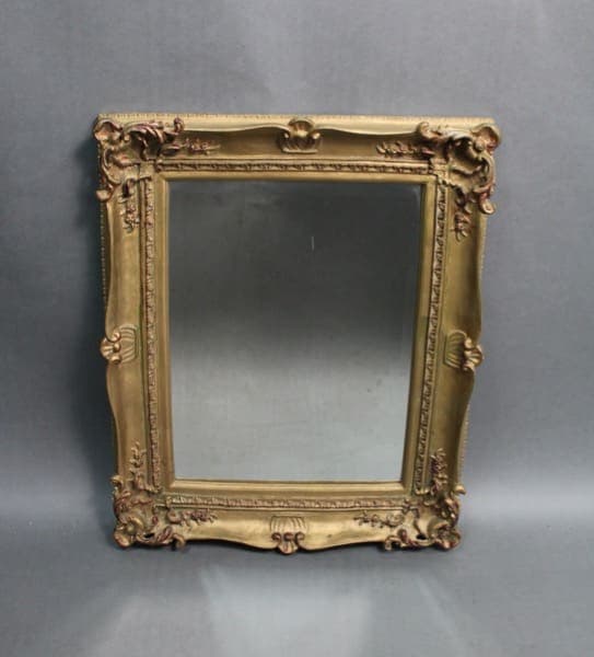 Antique ornate gold frame mirror with intricate carvings, featuring floral and scroll motifs, set against a plain grey background.