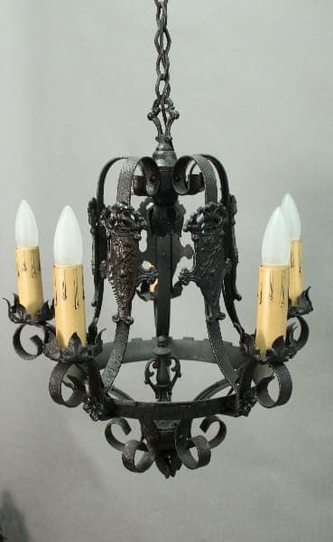 A 14408. Spanish Revival Chandelier With Shield Motif with six candle-shaped bulbs, featuring ornate metalwork and hanging from a chain against a gray backdrop.