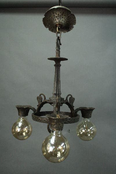 14601. Spanish Revival Three Light Chandelier with three lit bulbs, ornate metalwork, hanging from a ceiling mount in a gray room.