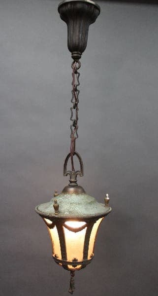 Turn Of The Century Pendant With Original Glass hanging lantern with a lit bulb, suspended by a chain from a dark ceiling mount, against a grey background.