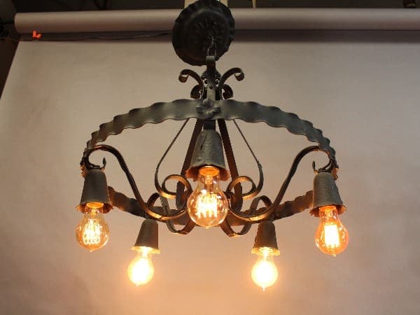 15370. 1920's Spanish Revival Chandelier with five glowing edison bulbs, mounted on a ceiling with a plain backdrop.
