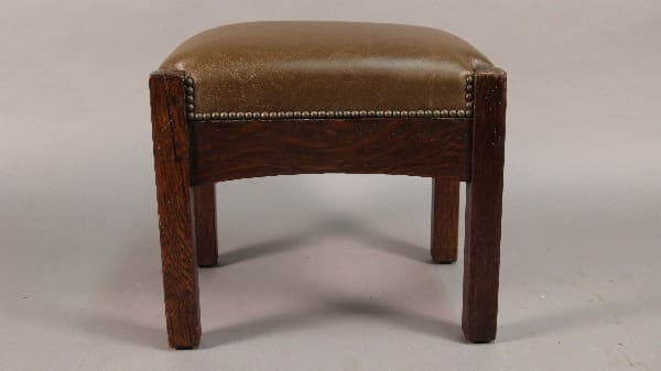 A small, vintage Antique Mission Arts And Crafts Arched Footstool with a cushioned, gold-toned seat and decorative metal studs along the edges.