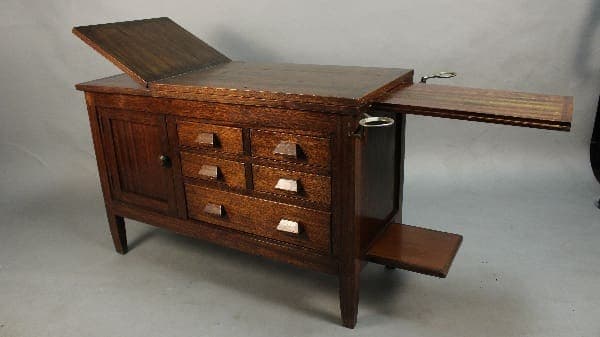 11436. Circa 1910 Arts And Crafts Oak Medical Examination Table with an open lid and pull-out writing surface, featuring multiple drawers and brass handles.