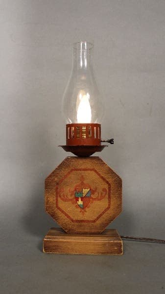 An antique 11724. 1930's Monterey Period Table Lamp with a lit wick, featuring a hexagonal wooden base and a delicate glass chimney, on a plain grey background.