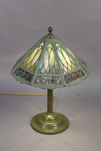 11754. Bradley and Hubbard Signed stained glass table lamp with a brass base and a greenish shade featuring intricate cut-out designs, displayed against a gray background.