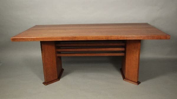 A 11986. Arts And Crafts Prairie Style Period Dining Room Table with a smooth top and panel legs, featuring a set of narrow shelves nestled underneath.