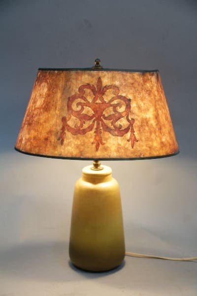 A table lamp with a yellow base and an aged paper shade featuring a red and brown floral design, illuminated in a dimly lit room. Product Name: 12021. School of North Dakota Lamp With Beautiful Period Mica Shade