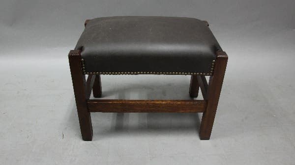 A small, Arts And Crafts 1910 footstool with new leather upholstery and brass stud detailing, positioned centrally against a grey background.