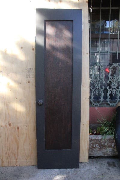 A modern narrow dark brown 14774. Spanish Revival Side Door #11 set in a plywood wall, with wrought iron decorative panels visible to the right.