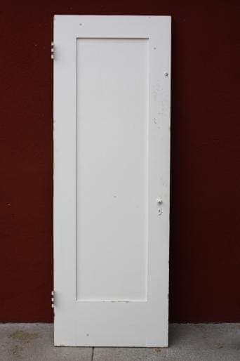 A white 6552. 1920s Single Panel Interior Door DD with a vertical rectangular panel, standing against a red wall, viewed from the front.